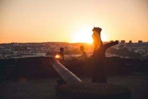 Silhouetted people at sunset with cityscape and cannon.