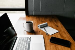 Laptop, coffee, notebook, and phone on wooden desk.