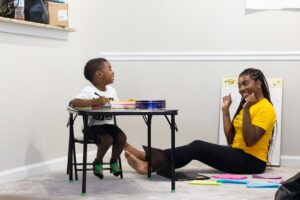 Child coloring with smiling woman sitting on floor.