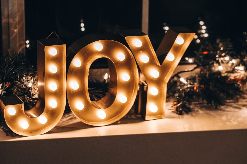 Illuminated 'JOY' sign with Christmas decorations in background.