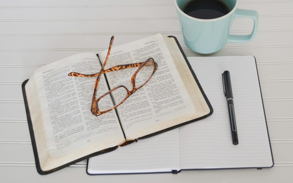 Open book, glasses, notebook, and coffee mug on table.