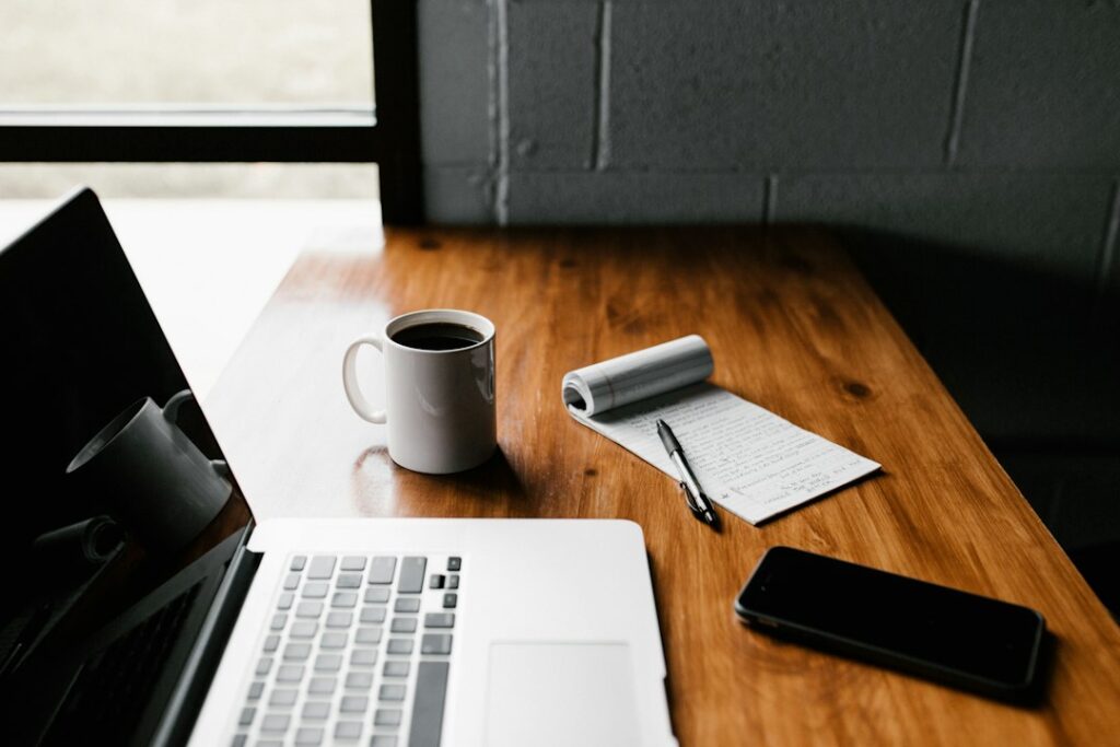 Laptop, coffee, notepad on wooden desk near window
