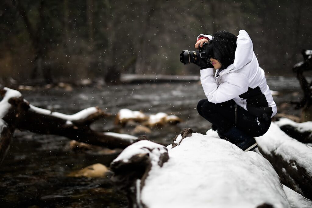 Photographer taking pictures in snowy forest beside river.