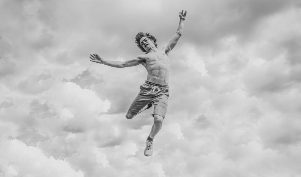 Man joyfully jumping against cloudy sky, black and white.